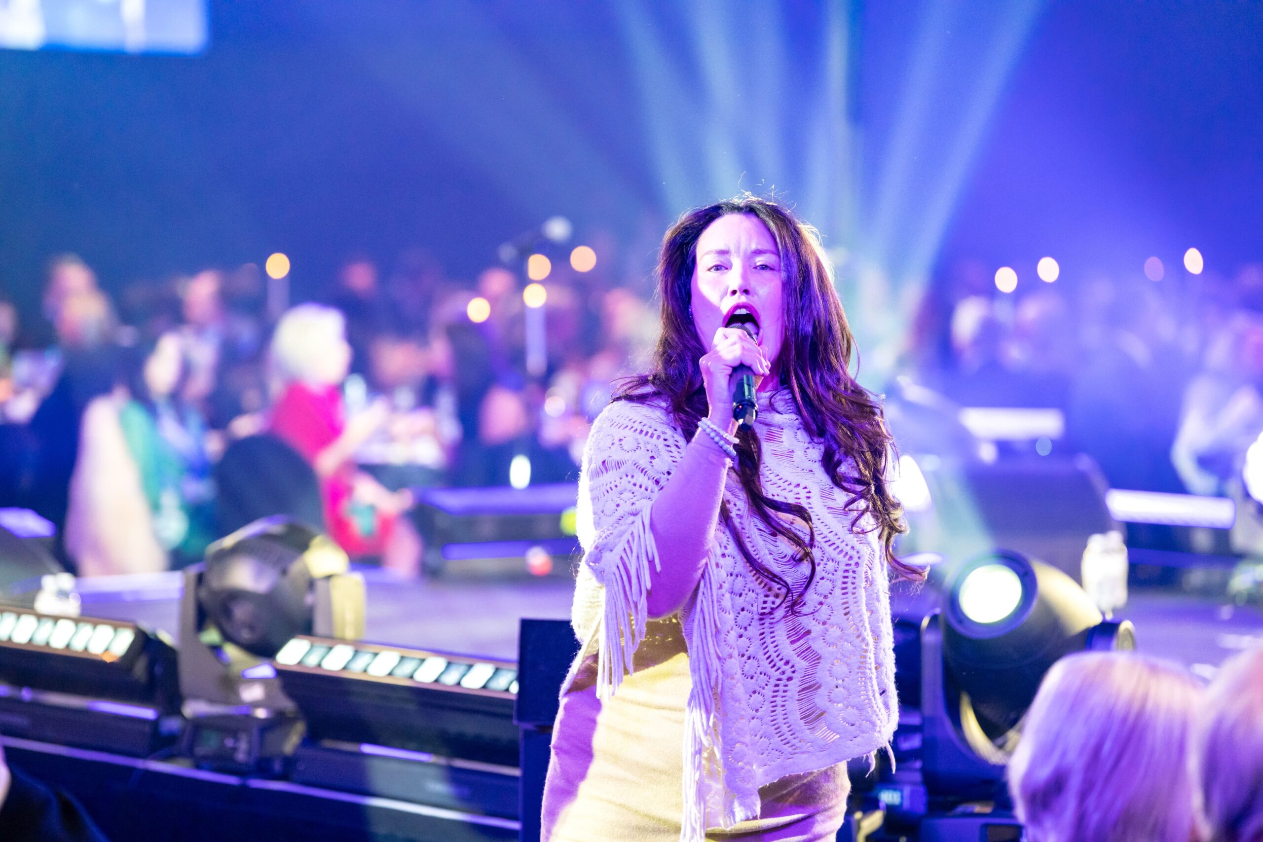 A woman with long hair sings passionately into a microphone on stage at Million Dollary Lunch surrounded by colorful lights and an audience seated at tables in the background.