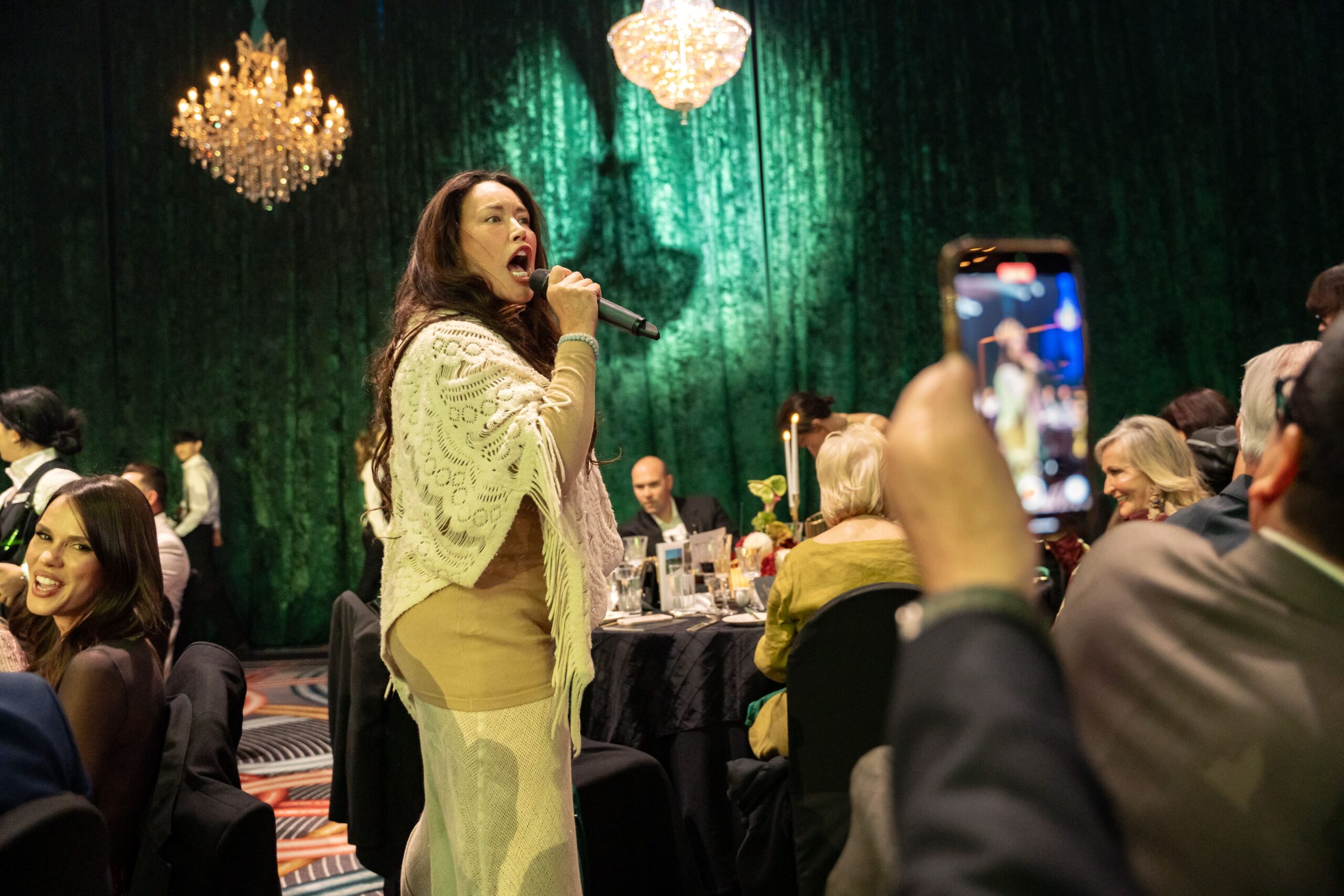 At ADIA 2023, a woman sings into a microphone among seated guests at a formal event. Chandeliers shine above, green drapes frame the room, and candlelit tables glow as someone in the foreground captures the moment on a phone.