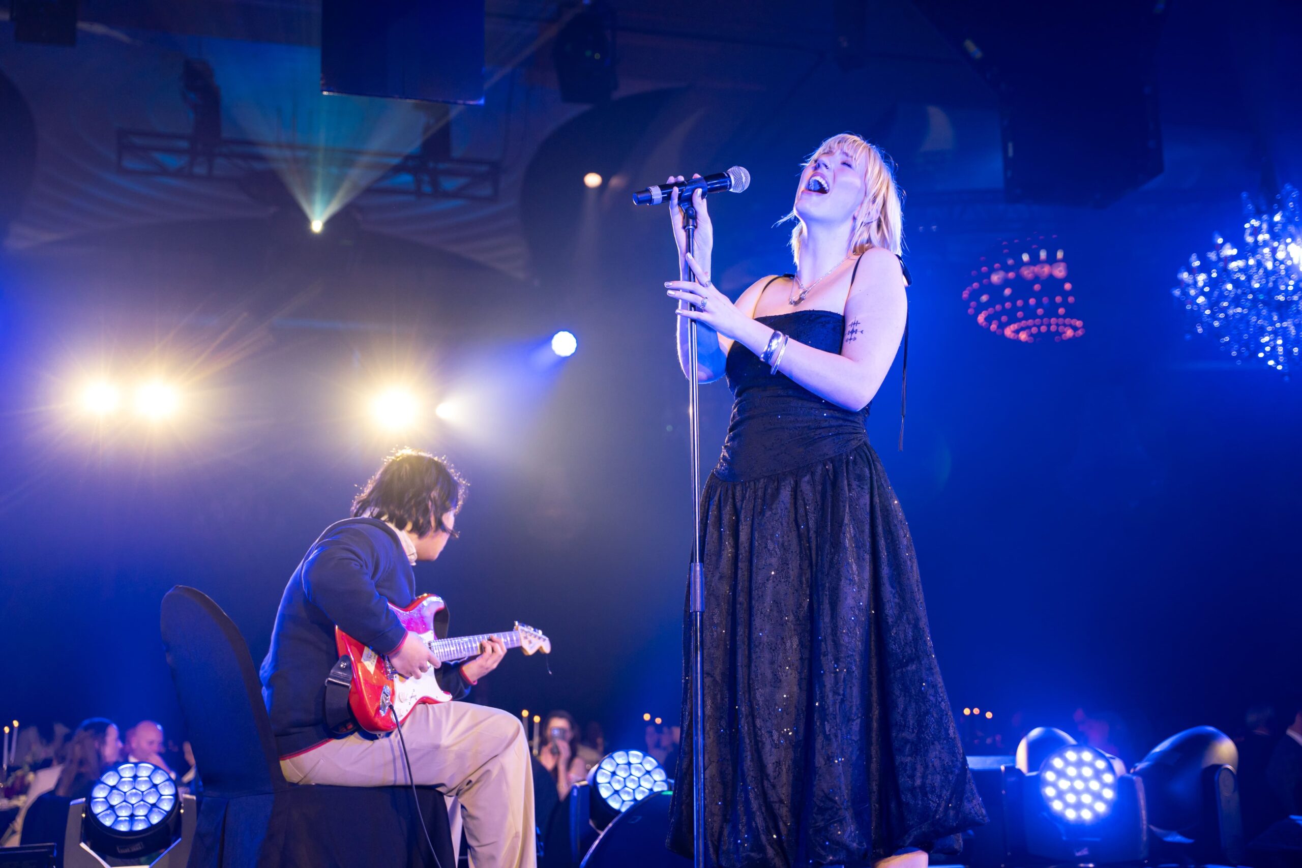 A woman in a black dress sings passionately into a microphone on stage at Million Dollar Lunch, while a seated man plays an electric guitar beside her. Bright stage lights and an enthusiastic audience are visible in the background.