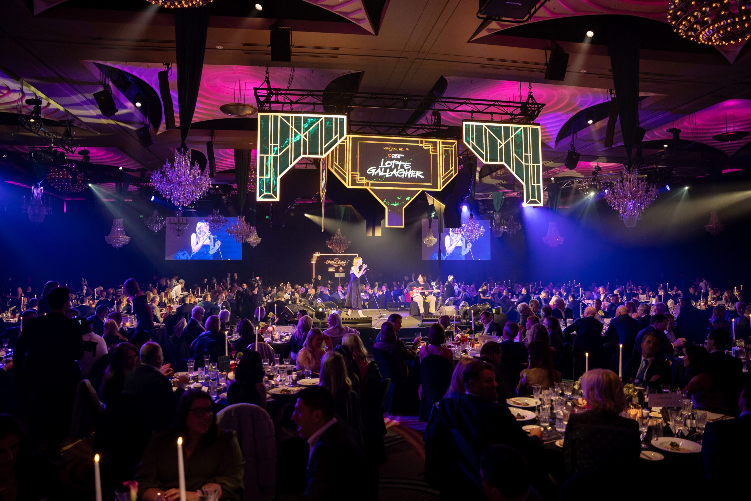 A large, elegant banquet hall full of seated guests, dramatic purple and yellow lighting, chandeliers, and a stage with performers. A large screen above the stage displays text reading “Lights, Glamour.” for the ADIA 2023 event.