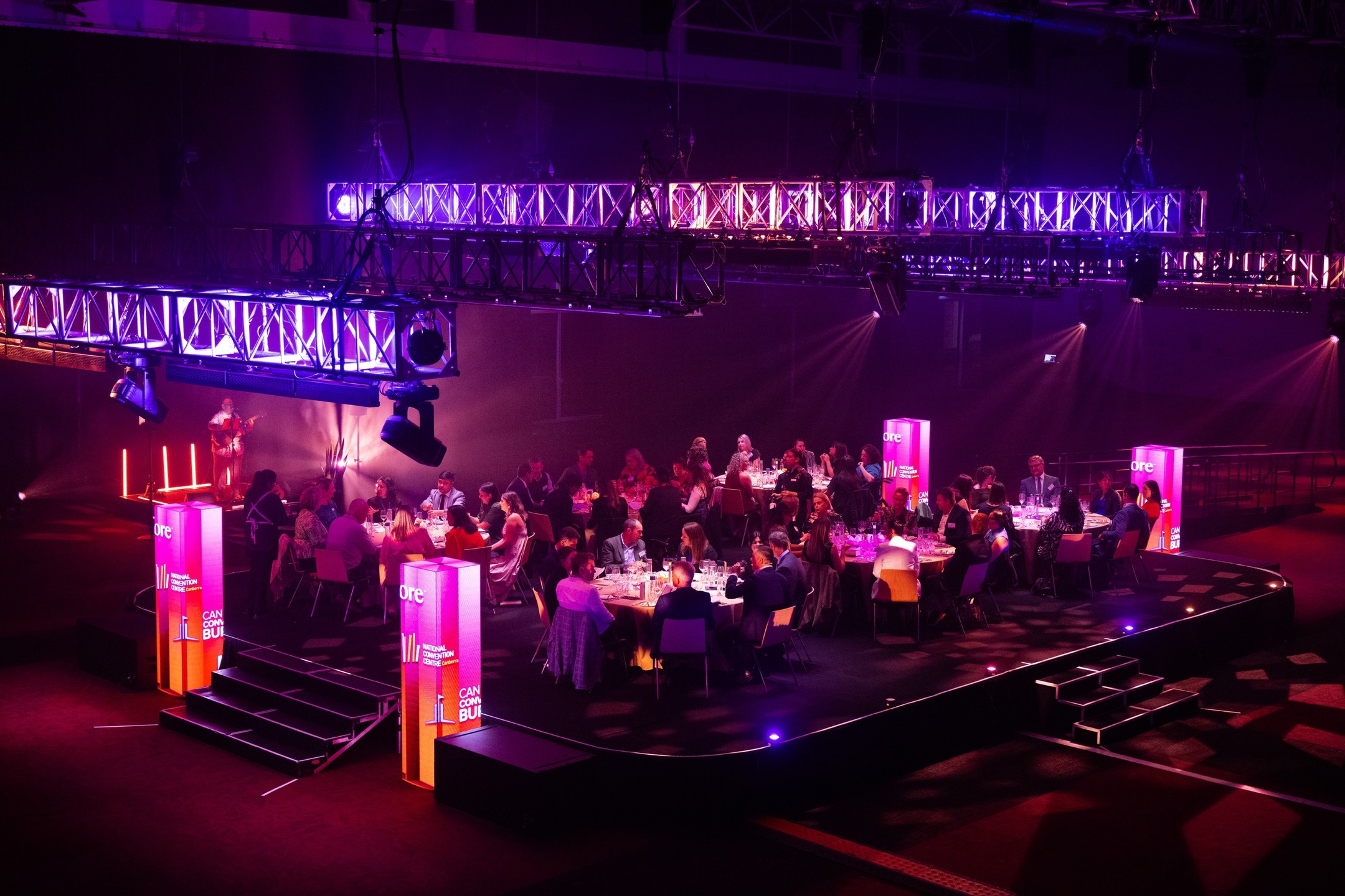 A group of people sit around tables at a formal event under dramatic purple and pink lighting, with large illuminated panels and stage equipment overhead in a dark, spacious venue.