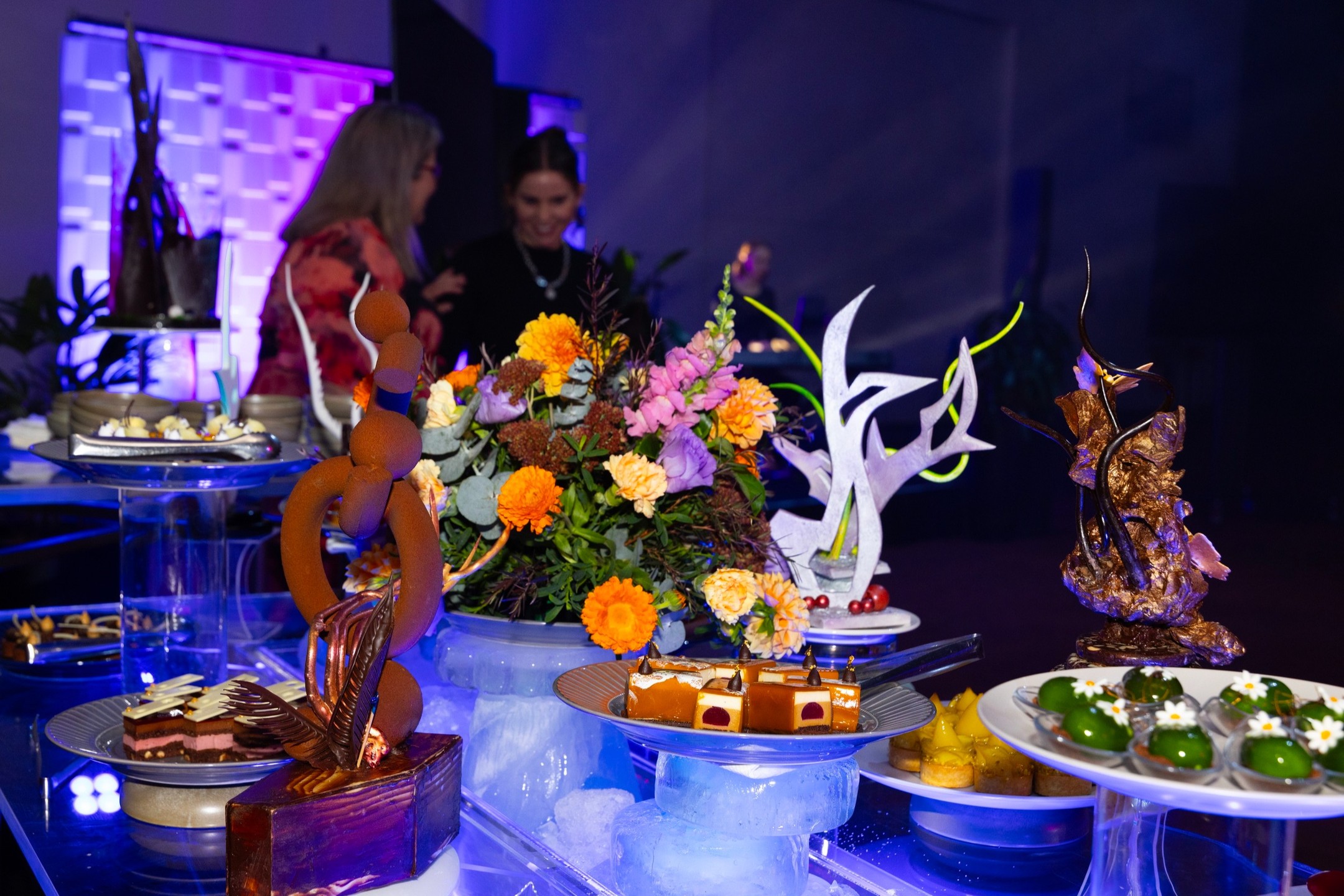 A display of elaborate desserts and chocolate sculptures on glass stands, with vibrant flower arrangements and two people conversing in the blurred background under purple lighting.