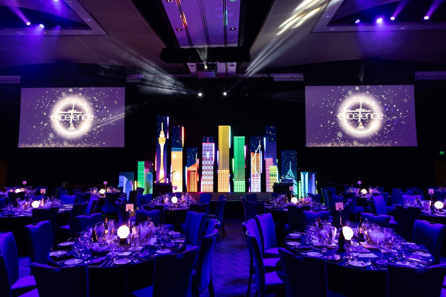 A banquet hall decorated with round tables set for dinner, glowing purple lights, and a stage featuring colorful skyscraper props. Two large screens display the word defence with a circular logo.
