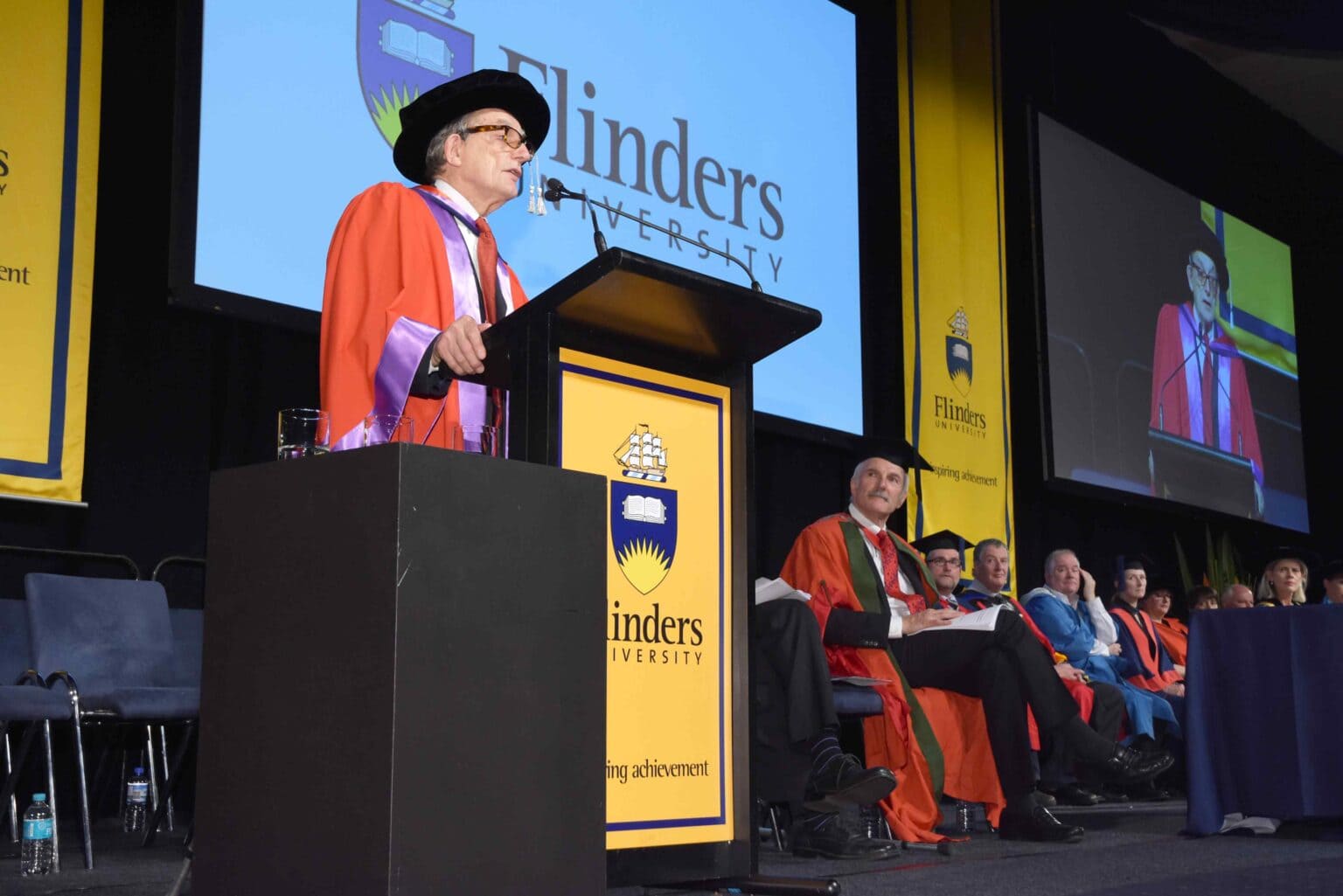 A person in academic regalia speaks at a podium during a Flinders University graduation ceremony, with faculty members seated on stage and the university logo displayed on banners and a screen.