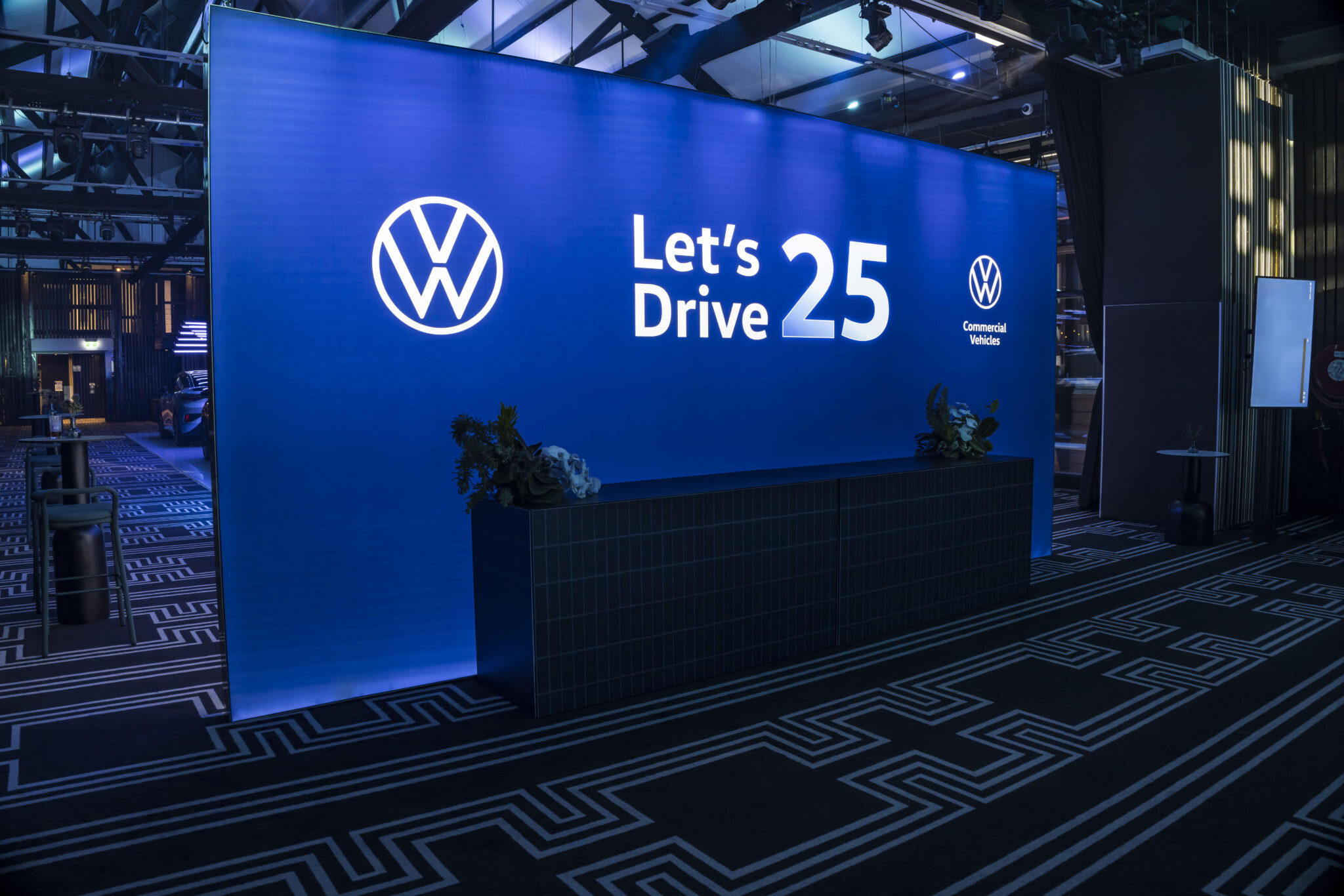 A large illuminated blue display with the Volkswagen logo reads Lets Drive 25 and Commercial Vehicles in a modern, dark room with patterned flooring. A black counter is placed in front of the display.