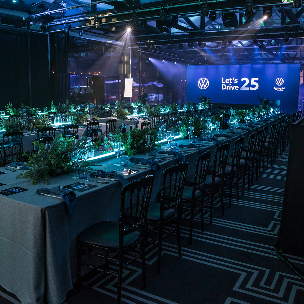 A formal event venue with long tables set for dinner, decorated with greenery and illuminated by blue lighting. A large screen at the front displays the Volkswagen logo and the text Lets Drive 25.