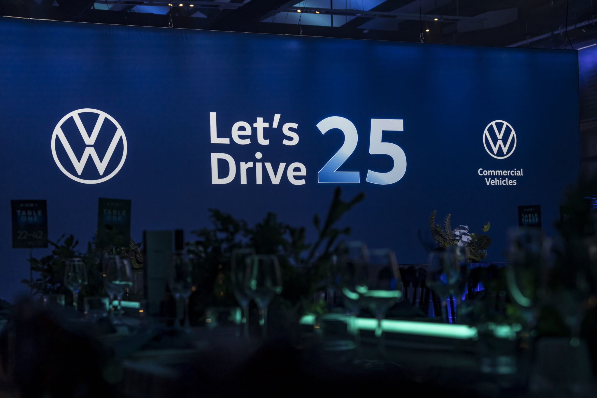 A large blue-lit backdrop displays the Volkswagen logo, the words Lets Drive 25, and Commercial Vehicles at an indoor event with tables and glassware in the foreground.