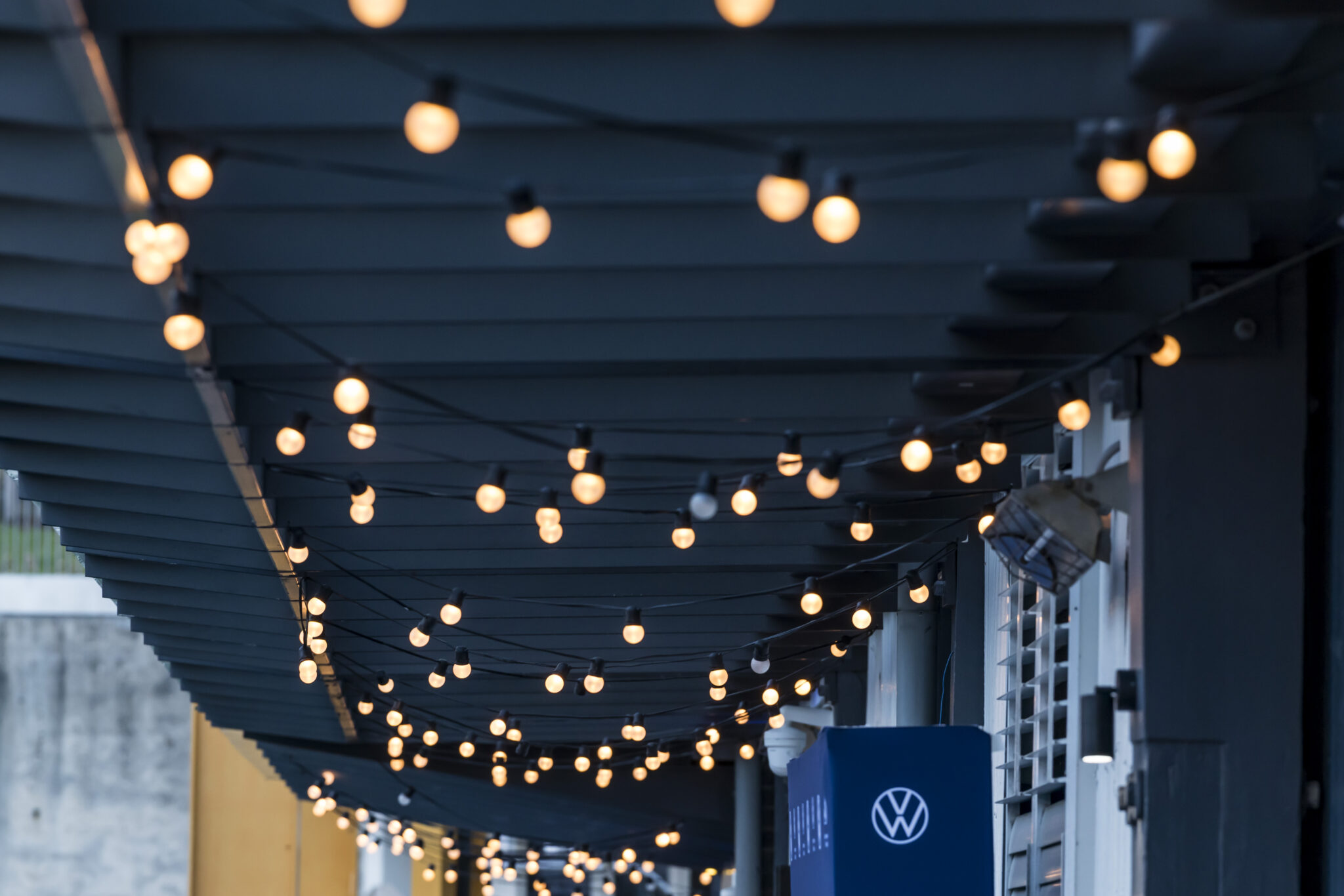 String lights with warm bulbs are hanging across a dark pergola, creating a cozy ambiance above an outdoor walkway. A Volkswagen sign is visible on the right side of the image.