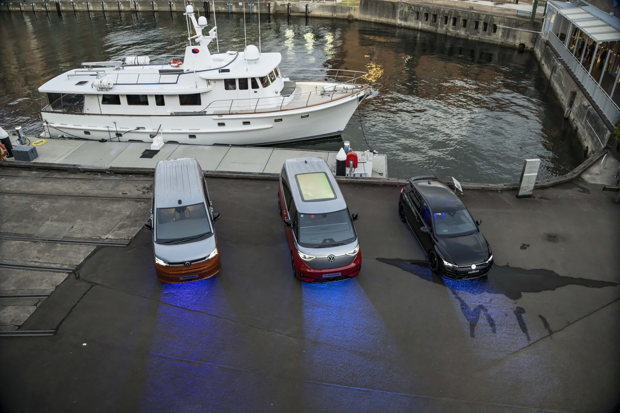 Three vehicles—a brown and white van, a red and white van, and a black car—are parked side by side near a waterfront, with blue lights shining underneath them and a white yacht docked in the background.