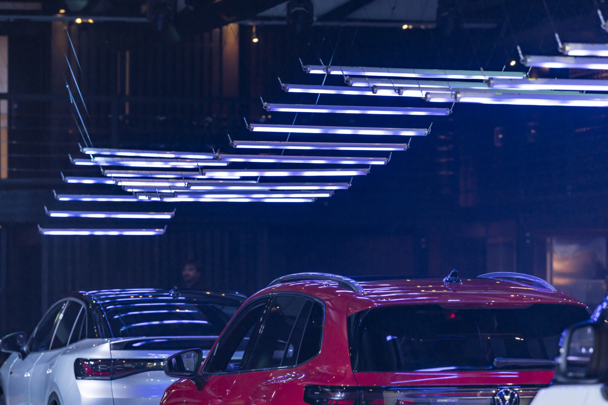 Two cars, one white and one red, are parked indoors under a series of modern, horizontal blue and white LED lights suspended from the ceiling. The scene appears to be part of a contemporary showroom or event space.