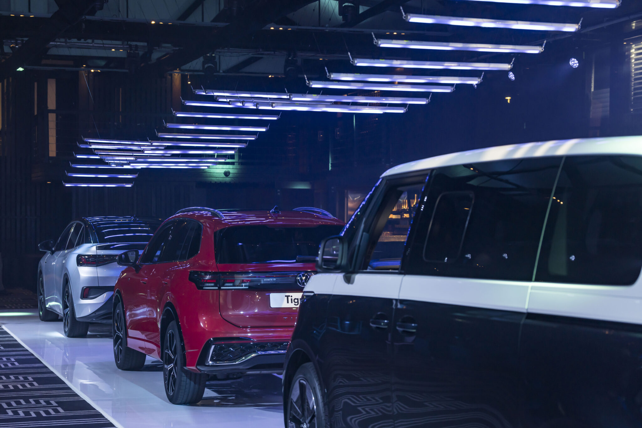 A row of three modern cars, including a red SUV and a two-tone black-and-white van, are displayed indoors under futuristic, hanging LED lights.