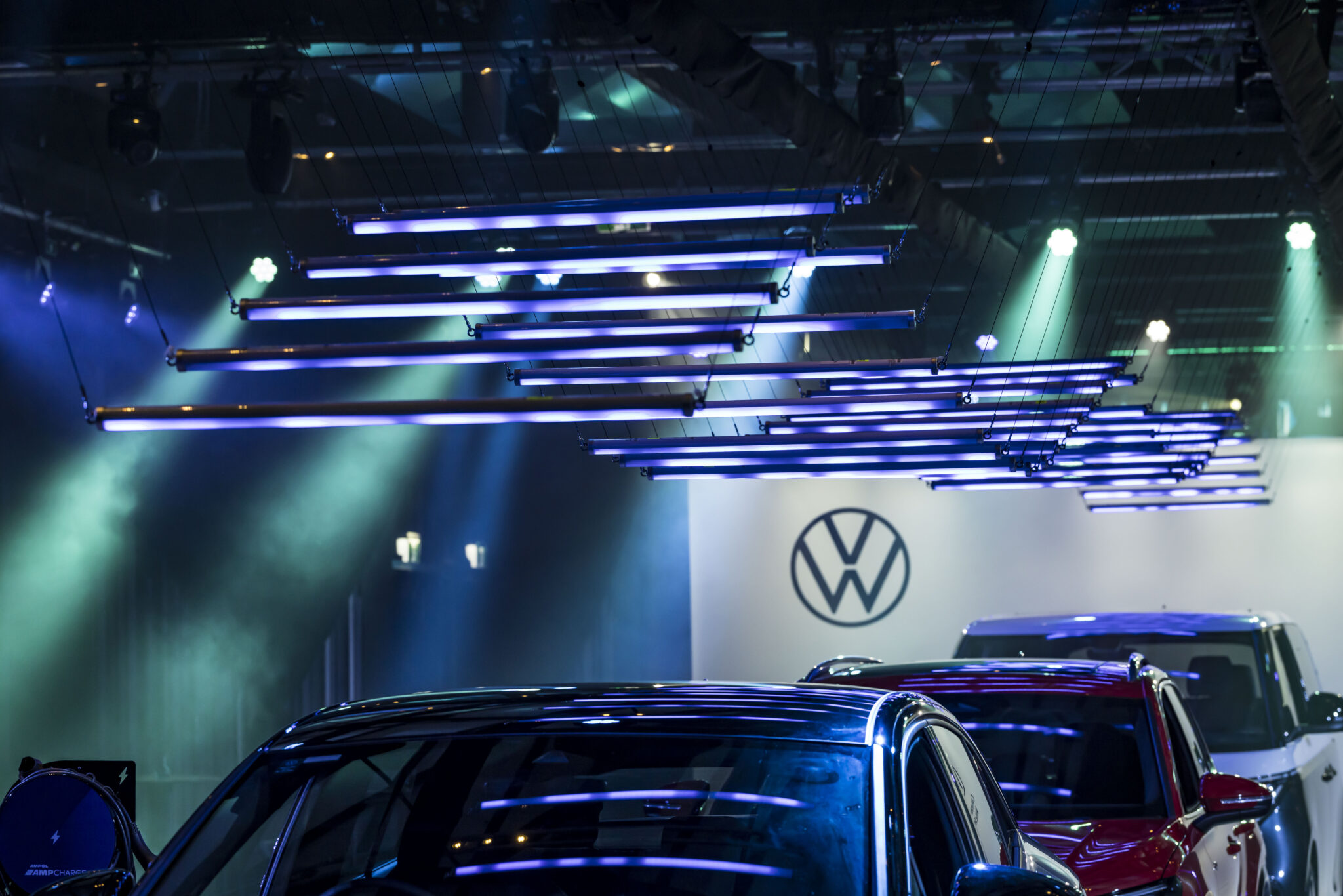 Rows of blue and purple LED lights hang above shiny cars at an indoor auto show. The Volkswagen logo is visible on a white wall in the background, illuminated by spotlights and surrounded by a misty atmosphere.