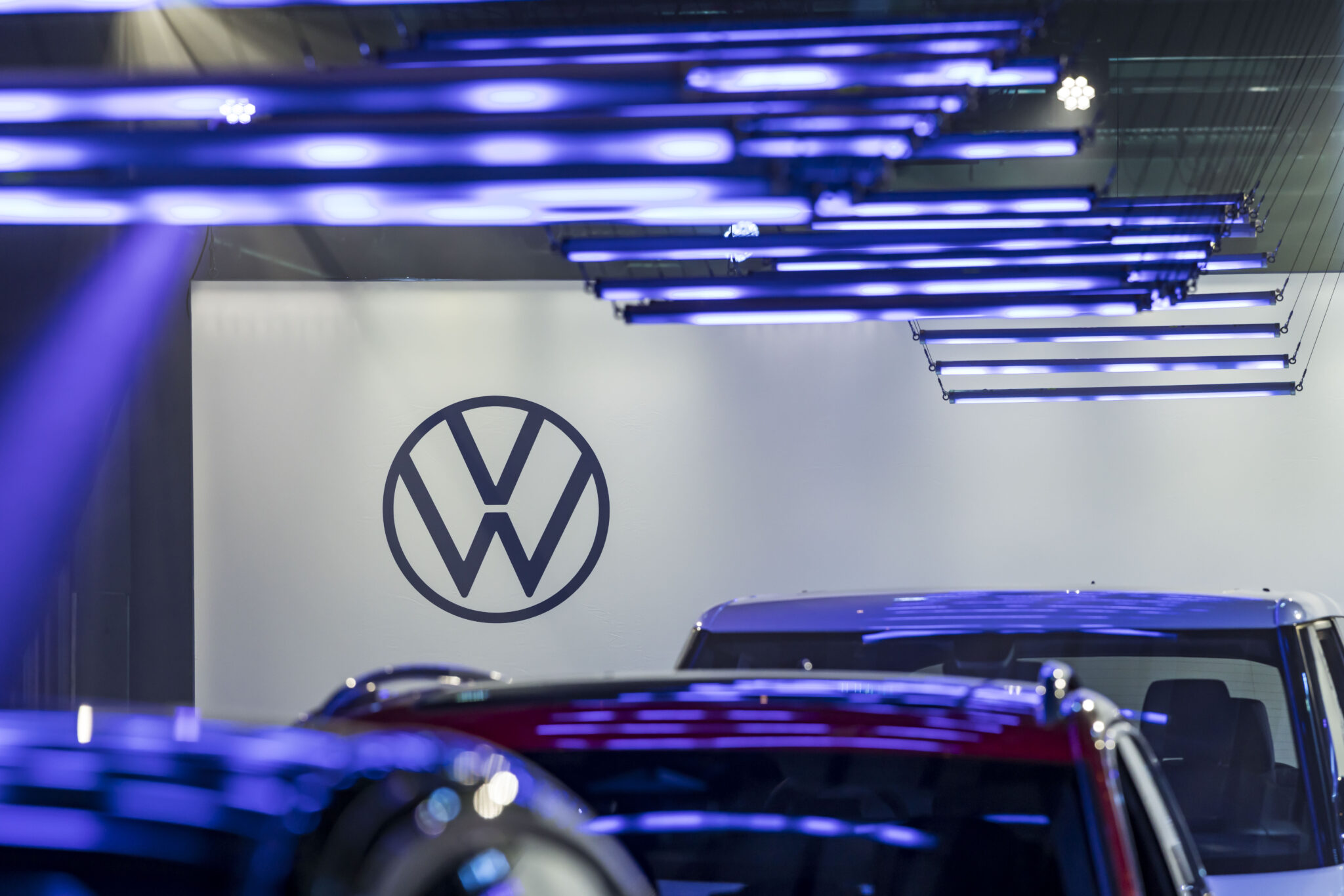 A Volkswagen logo is displayed on a white wall, with several cars and blue LED lights in the foreground at an auto show or showroom.