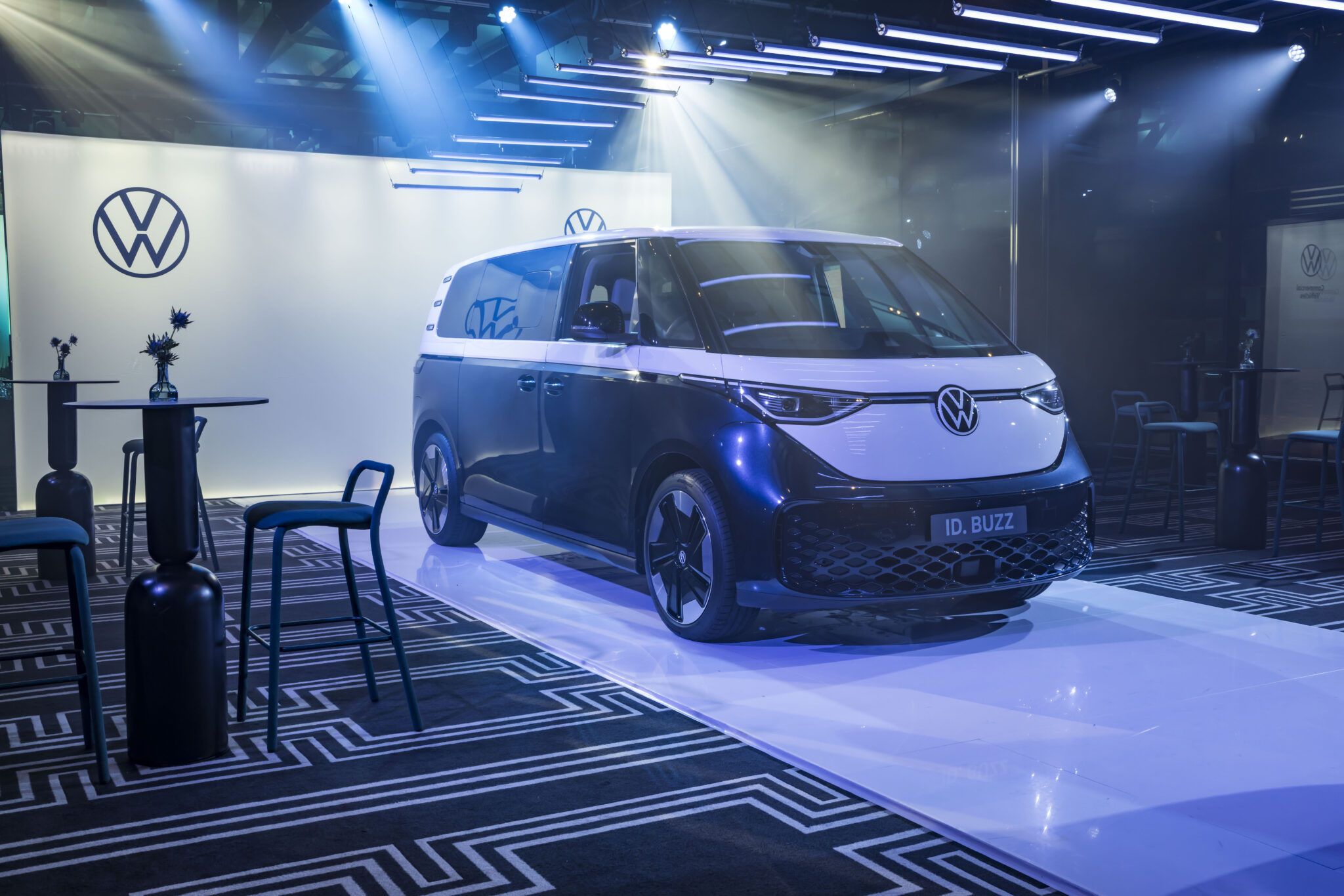 A blue and white Volkswagen ID. Buzz electric van is displayed on a glossy platform in a modern, stylish indoor showroom with geometric patterns, ambient lighting, and minimalist decor.