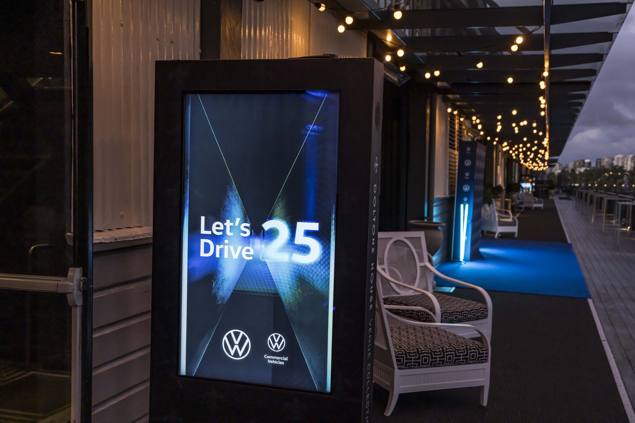 A digital sign displays Lets Drive 25 with Volkswagen and Commercial Vehicles logos, set on an outdoor deck with string lights, white chairs, and a blue carpet at dusk.