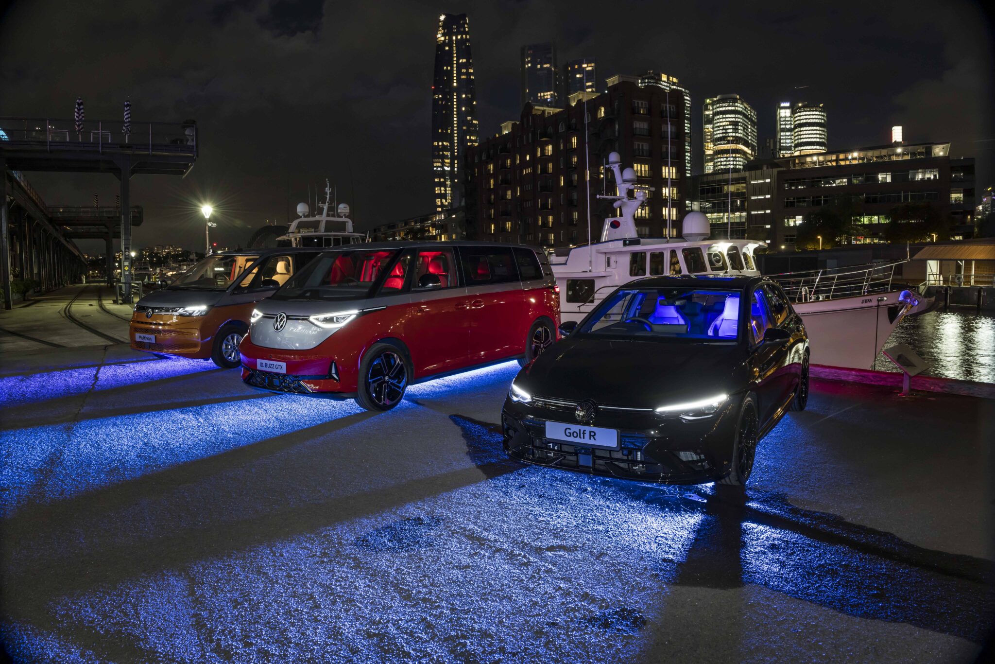 Three Volkswagen vehicles parked on a dock at night are illuminated by blue and purple lights, with modern buildings and yachts visible in the background.