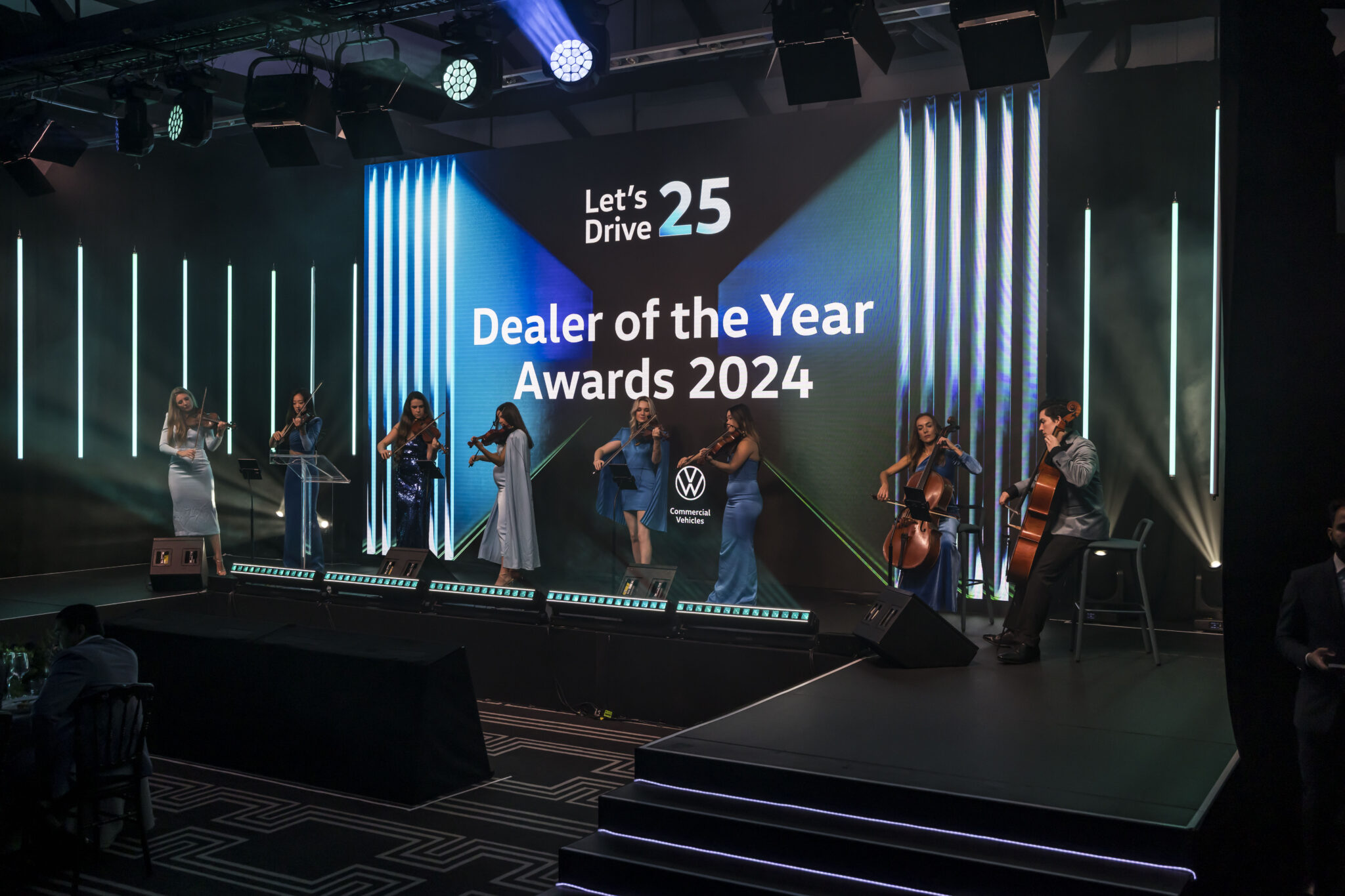 A string ensemble performs on stage at the Dealer of the Year Awards 2024 event, with blue lighting and a large screen displaying Lets Drive 25 and the Volkswagen Commercial Vehicles logo.