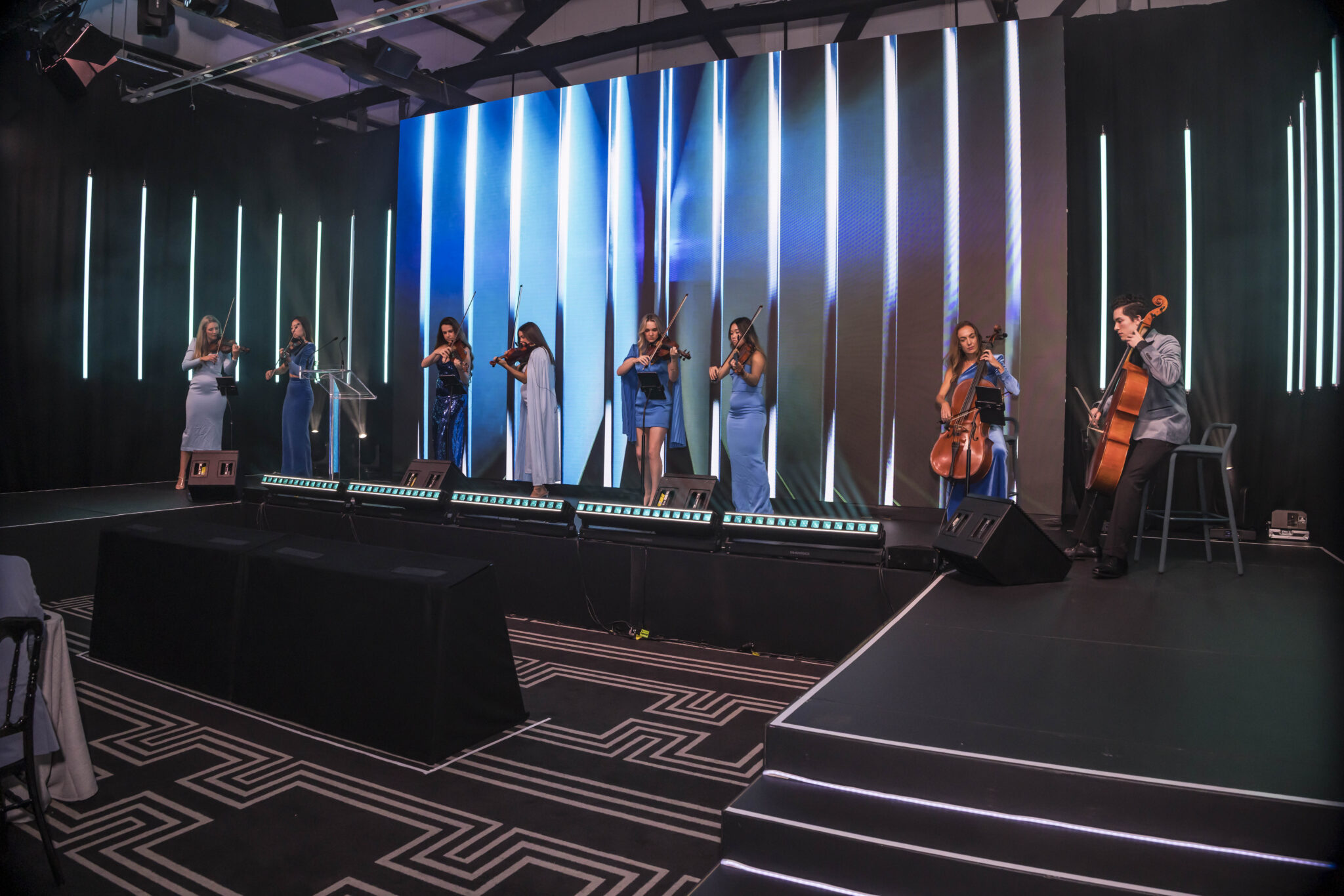 A group of musicians in blue dresses plays string instruments on a stage with vertical light bars and a large digital screen in the background. The venue has dark walls and patterned flooring.
