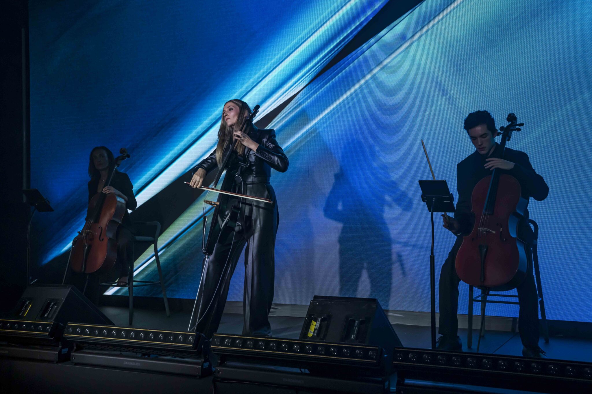 Three musicians in dark clothing play cellos onstage, illuminated by blue and green stage lights and abstract background visuals. The central musician stands, while the others sit on either side.
