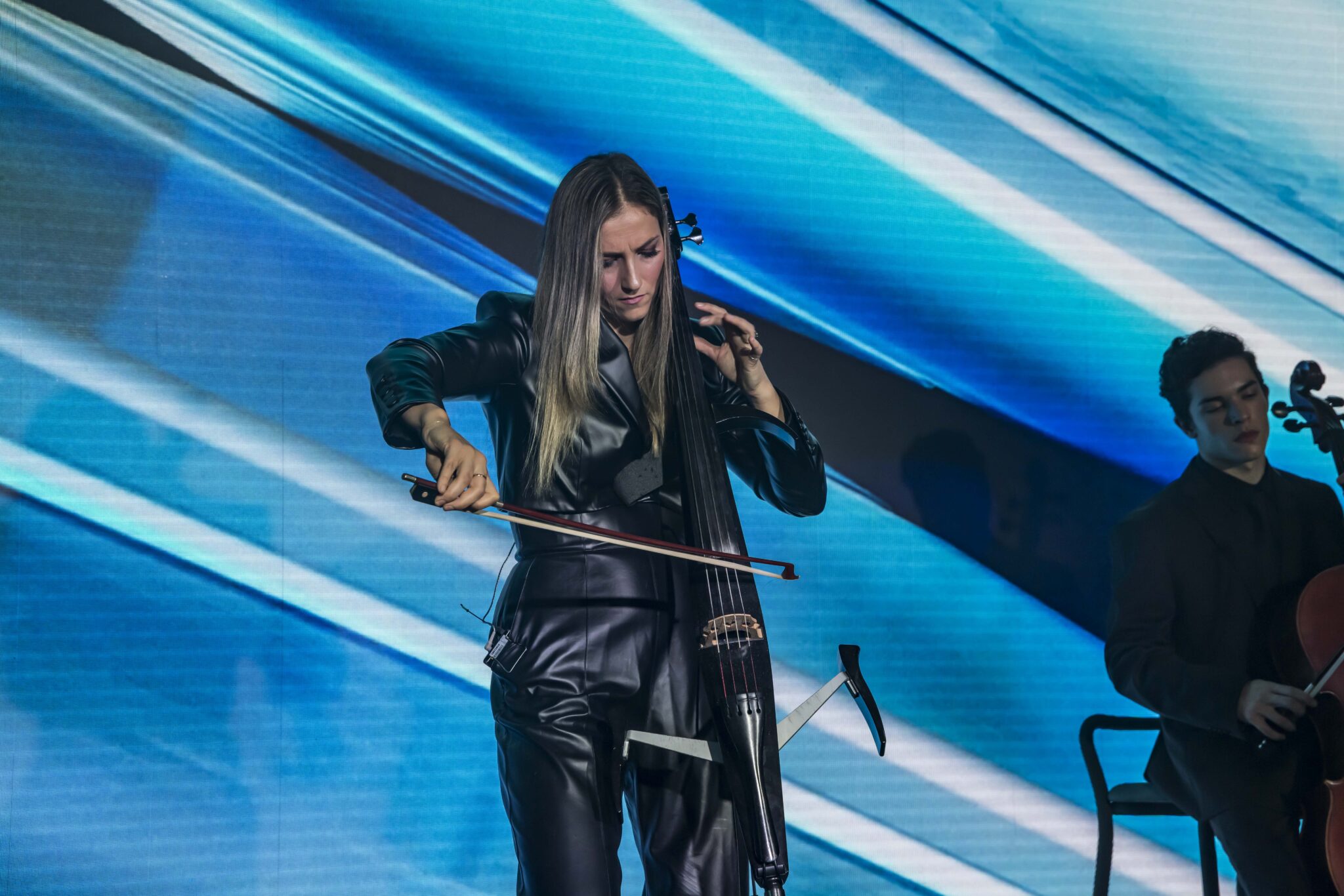 A woman in black leather clothing plays an electric cello on stage with a blue abstract background. Another musician is partially visible, seated and playing a cello.