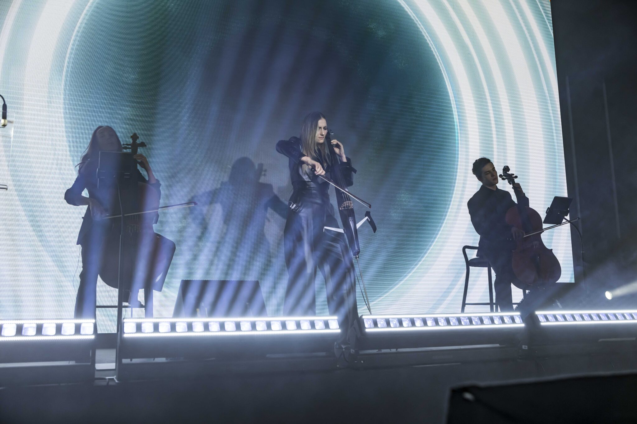 Three musicians play string instruments on stage, illuminated by dramatic lighting and a circular light pattern on a screen behind them, creating a vibrant and atmospheric concert scene.