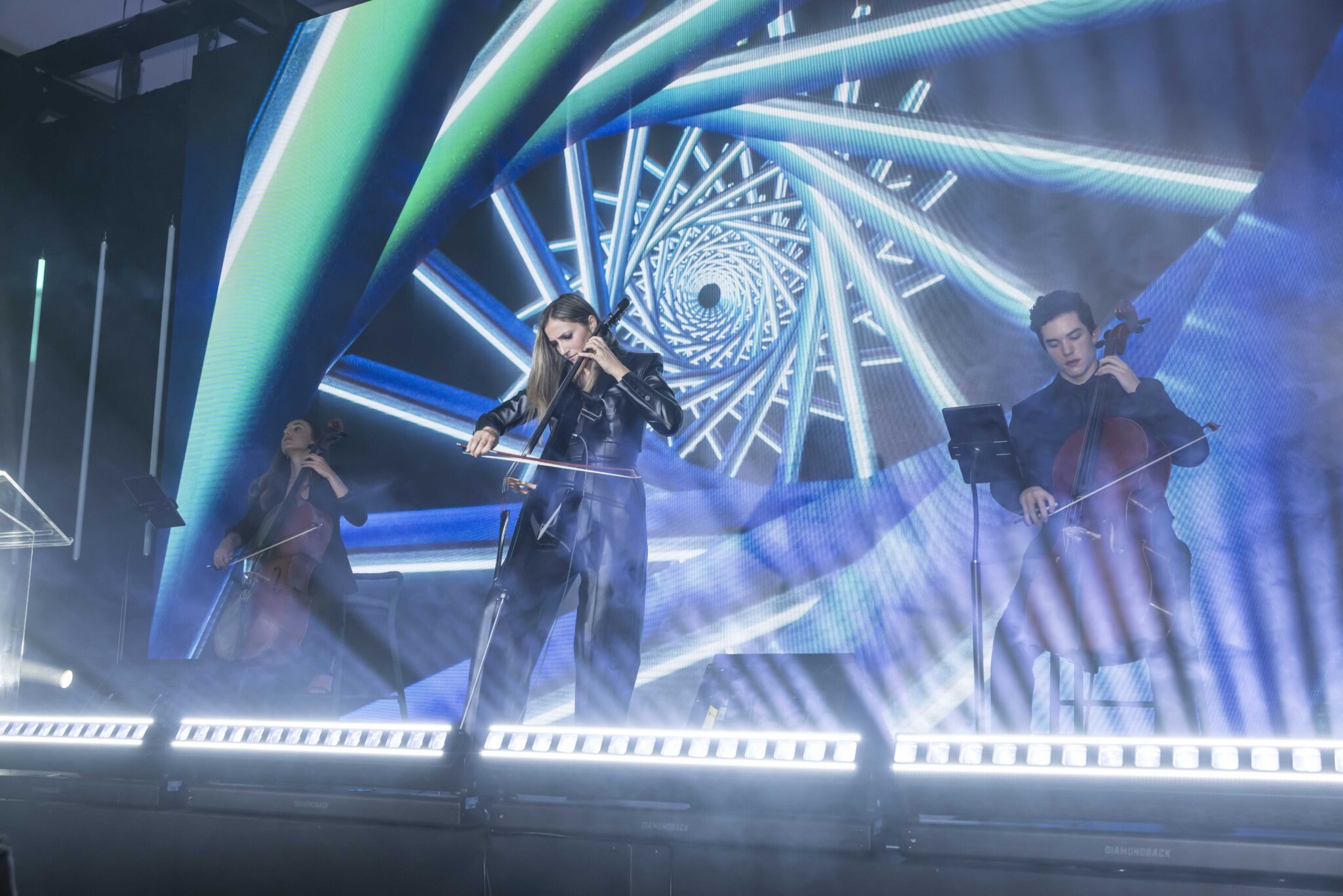 Three musicians play string instruments on a brightly lit stage, with a swirling blue and green spiral pattern displayed on a large screen behind them, creating a dynamic and futuristic atmosphere.
