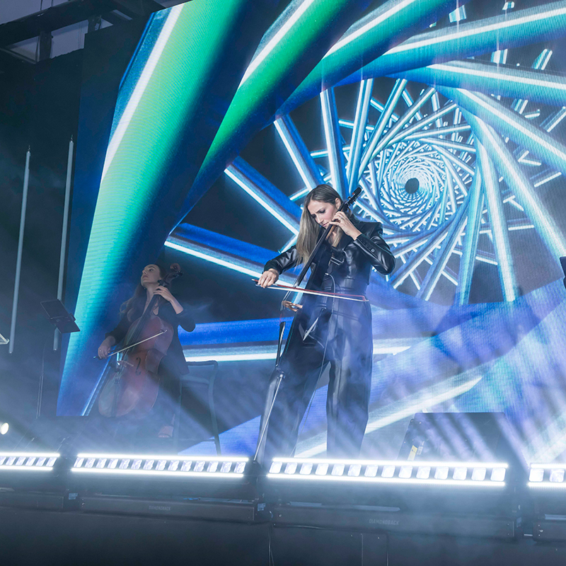 Two musicians play string instruments on a stage, one in the foreground with a cello. Behind them, a large digital screen displays a blue spiral pattern, and bright lights and mist create a dramatic atmosphere.