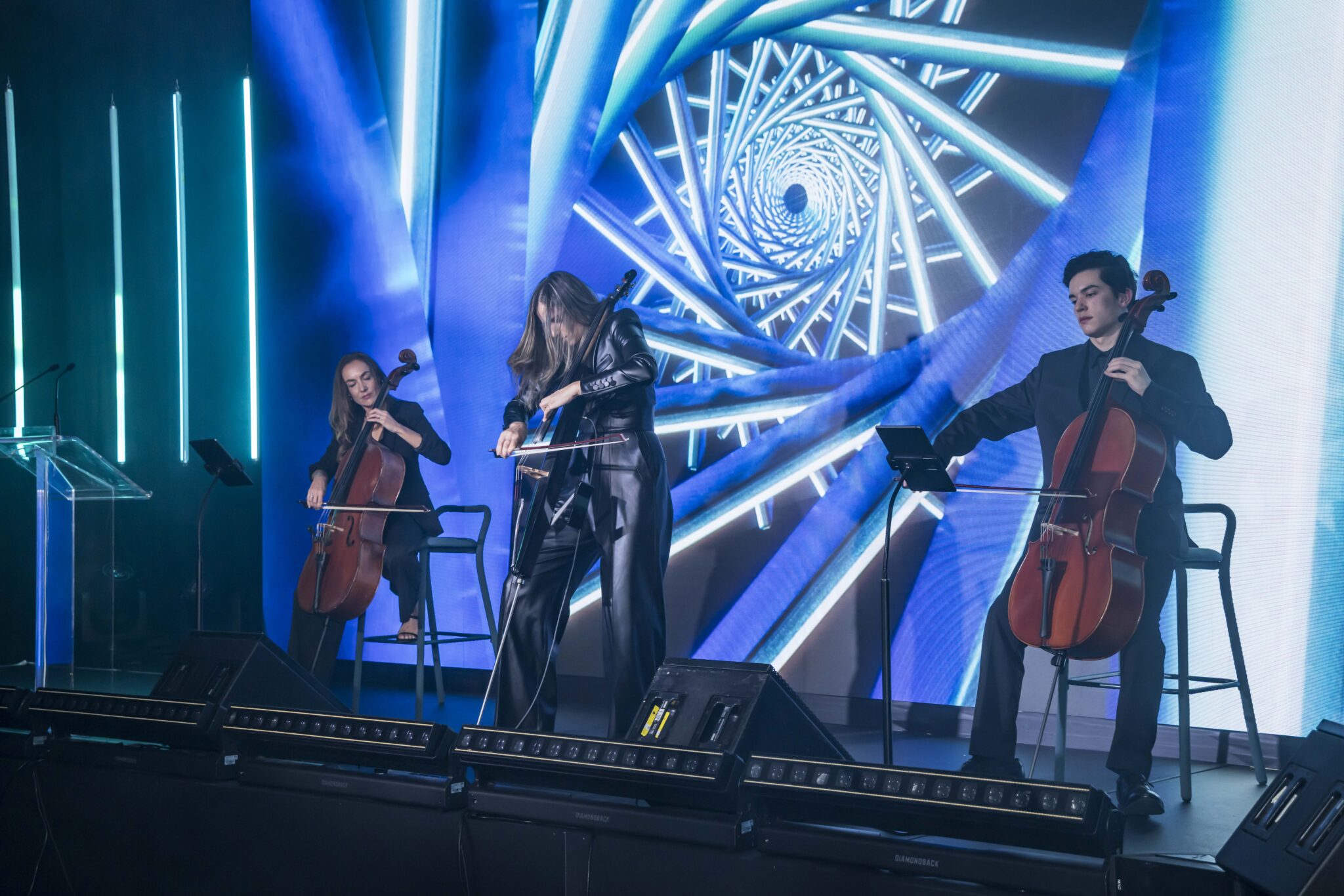 Three musicians play cellos on stage in front of a large, colorful digital screen displaying a blue spiral pattern. They are dressed in black and illuminated by stage lights.
