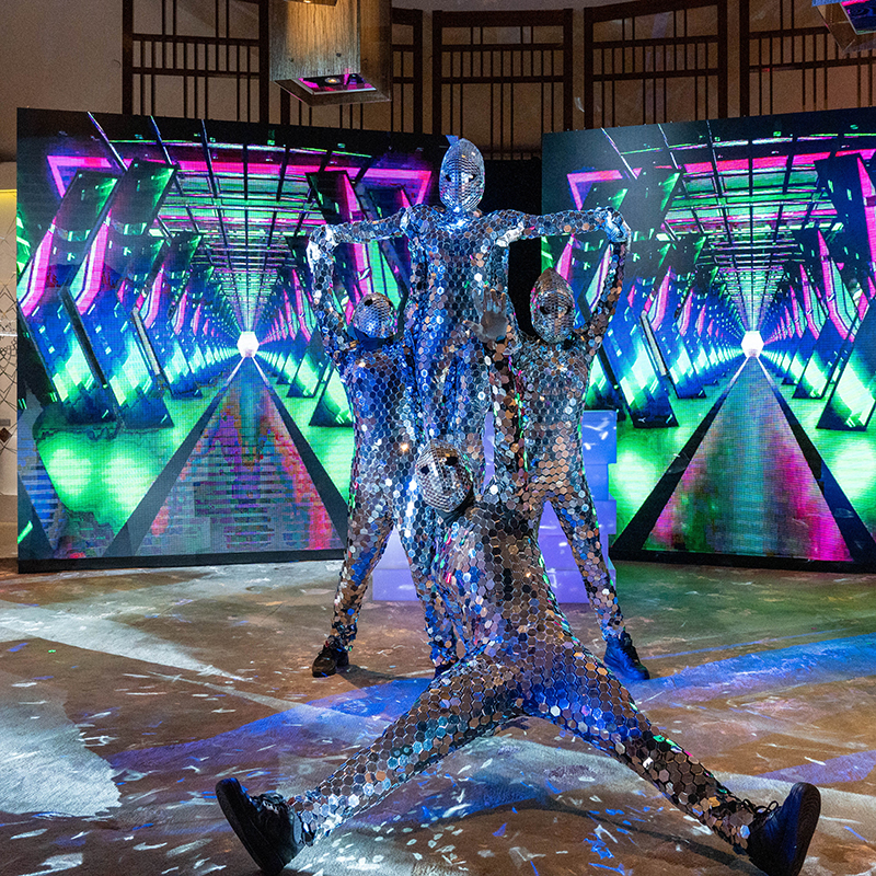 Four performers in reflective, mirror-like suits pose before vibrant digital screens displaying a futuristic tunnel—an electrifying scene by a leading event production company in Singapore, blending striking visuals with energetic movement.