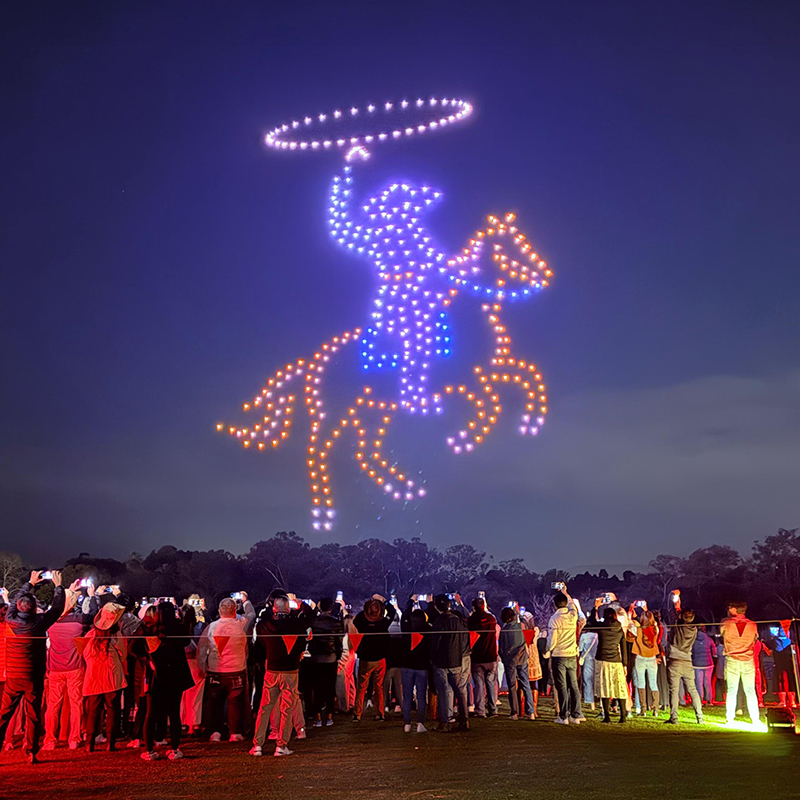 A crowd watches a nighttime drone light show, produced by a leading Perth event company, as glowing drones form a cowboy on horseback with a raised lasso against the night sky.