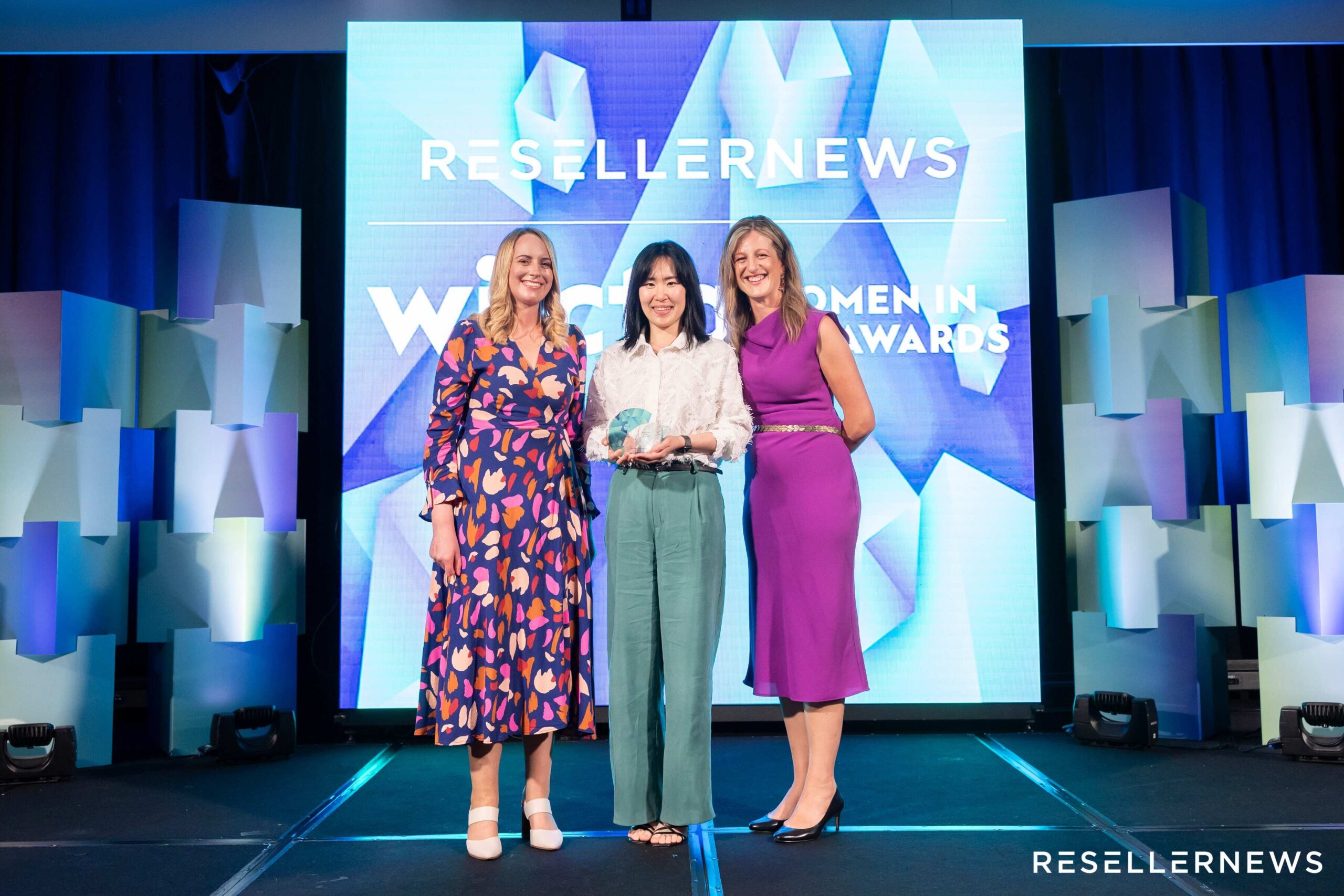 Three women stand smiling on stage at an awards event. The woman in the center holds a glass award. Behind them, a screen displays RESELLERNEWS Women in ICT Awards. The setting is formal with stage lighting.