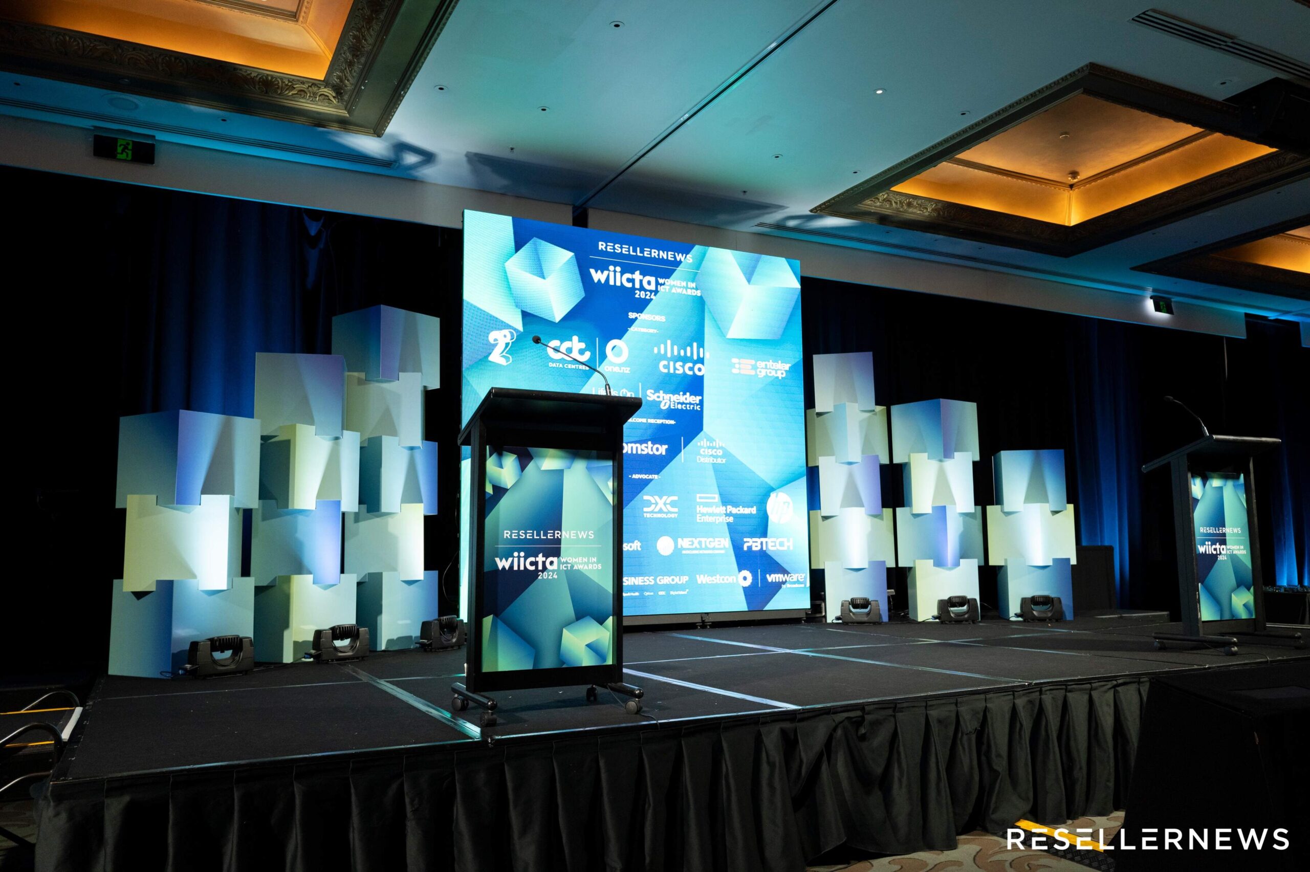 A conference stage with blue geometric displays, two podiums, and large screens showing sponsor logos including Cisco, Schneider Electric, and others. The event branding reads “Reseller News WIICTA.”.