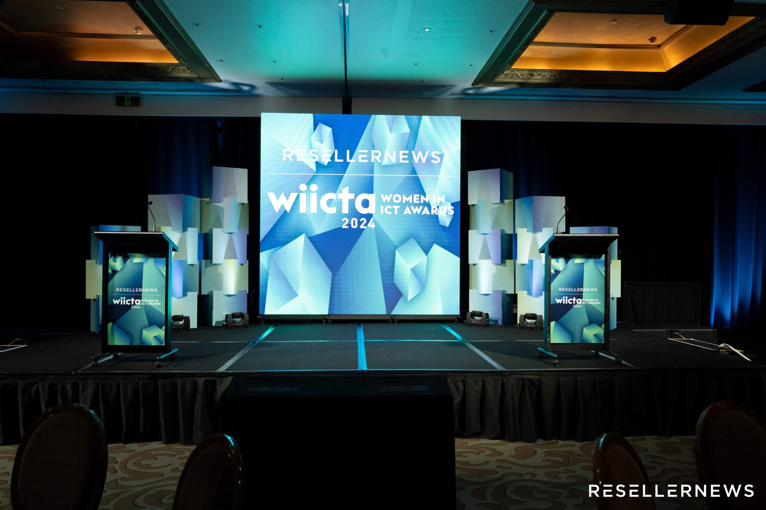 An empty stage setup for the 2024 Women in ICT Awards (WIICTA) with two podiums, geometric backdrop panels, large central screen displaying event branding, and Reseller News logo visible.