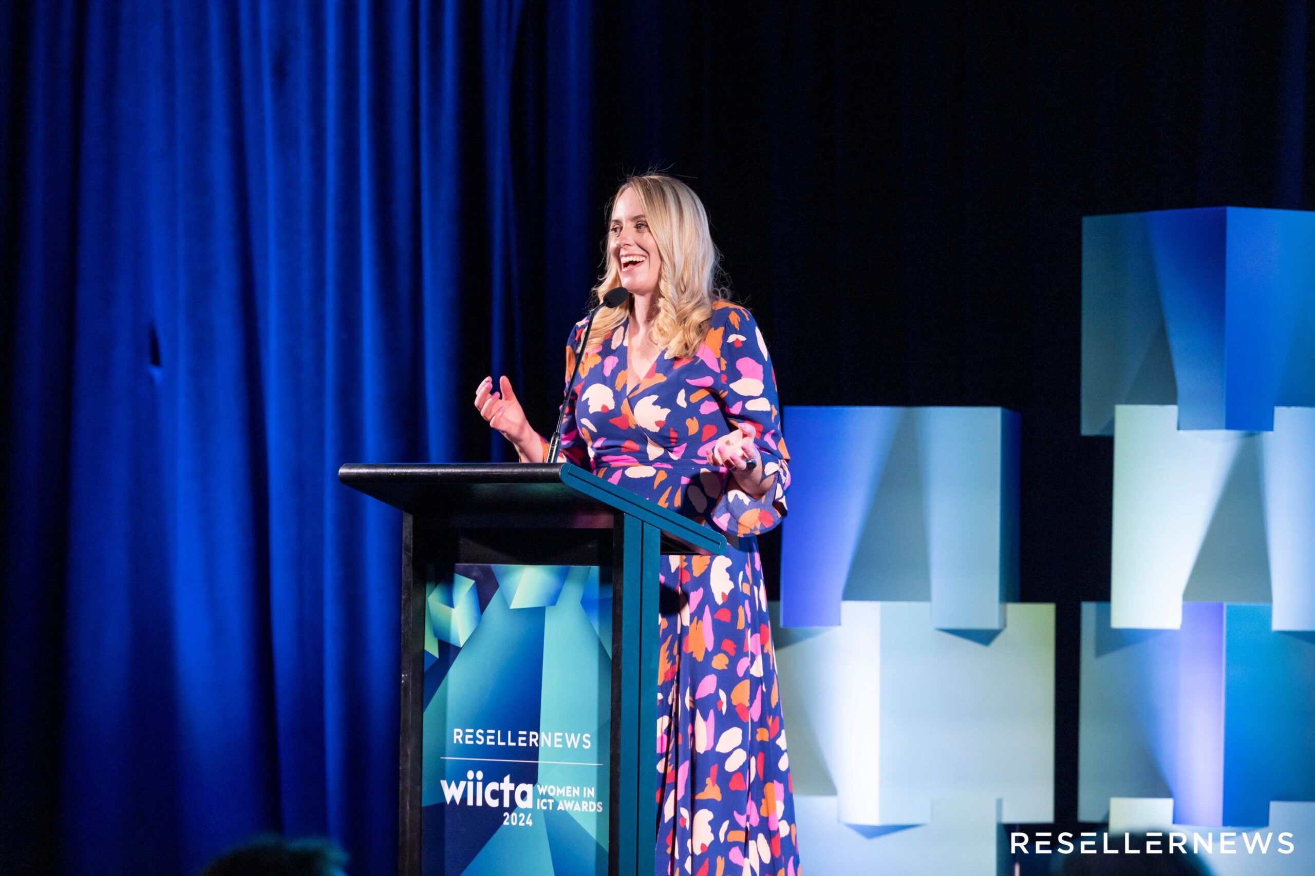 A woman in a colorful, long floral dress speaks at a podium on stage at the 2024 Reseller News WiCTA Women in ICT Awards event, with blue geometric decorations and curtains in the background.
