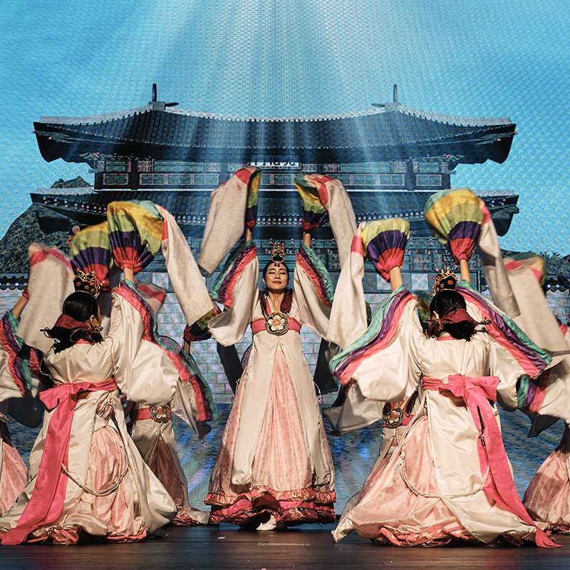 A group of women in traditional Korean dresses perform a fan dance on stage at an event production in Seoul, with colorful fans raised. A backdrop featuring a traditional Korean building is illuminated behind them.
