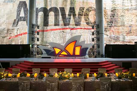 A stage set for a gala dinner features red steps and a centerpiece resembling the Sydney Opera House, with a backdrop displaying the partially visible word Amway. Tables adorned with candles and flowers are arranged in front.