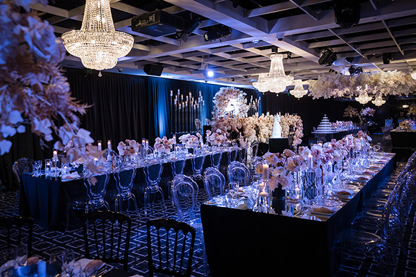 Elegant wedding reception hall perfect for weddings, with long tables set with glassware, candles, and floral centerpieces. Crystal chandeliers hang from the ceiling, and the room is softly lit with blue accents.