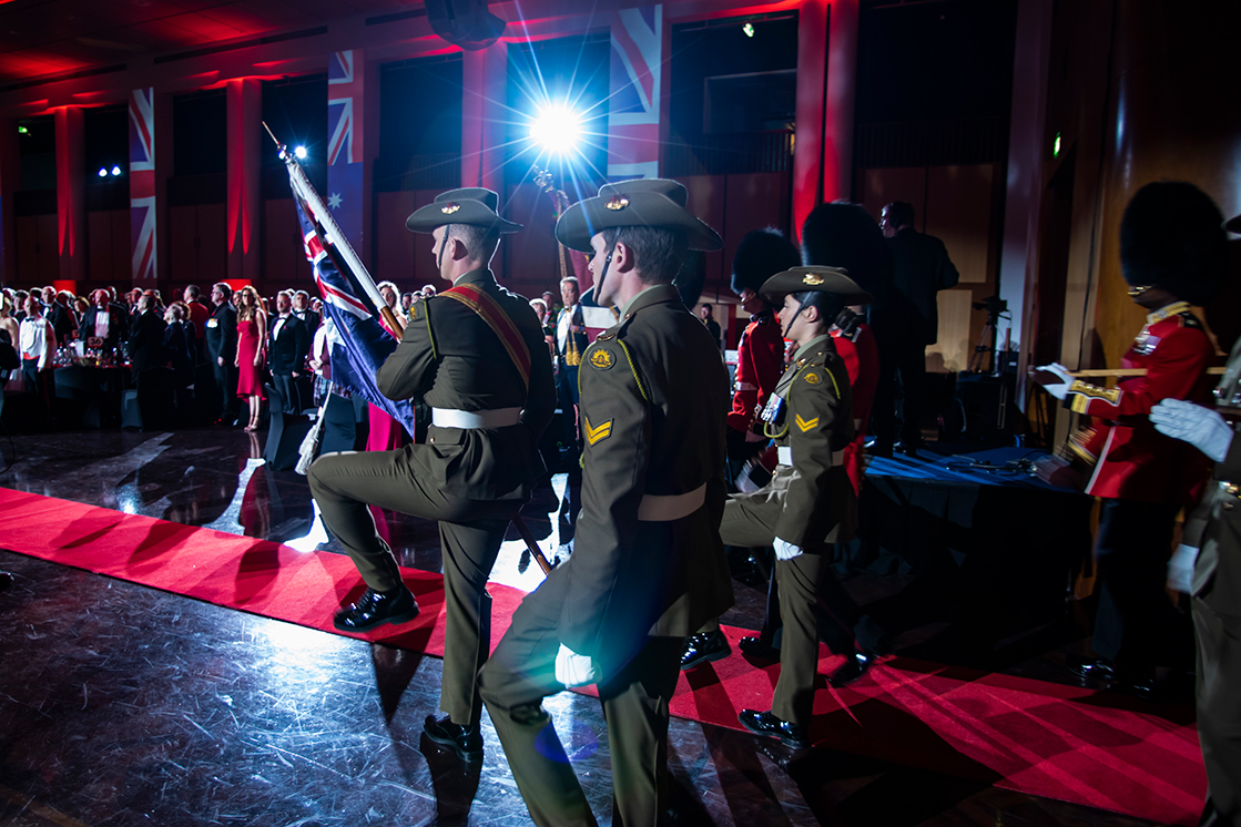 Uniformed soldiers march in formation on a red carpet indoors, carrying a flag. British flags hang in the background, and an audience stands to the side under dramatic lighting by Encore Canberra, an expert event production Canberra team.