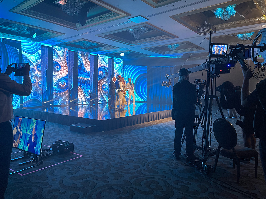 A stage with blue, abstract light panels features performers in costume. Camera operators and crew from an event production company in Auckland film the scene, with monitors and equipment visible in a dimly lit, ornate ballroom.