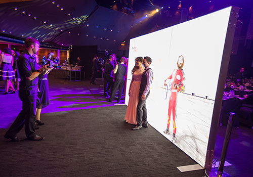 Three people pose in front of a large, brightly lit display screen—showcasing expert Designing Event Media Walls—at an indoor event. Two photographers capture the moment against purple lighting and background tables.