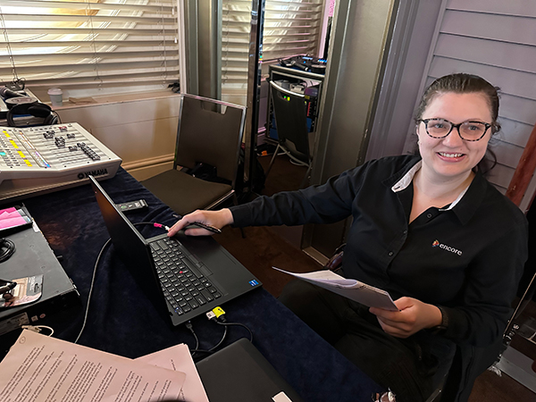A woman wearing glasses and a black shirt sits at a desk with papers, a laptop, and audio equipment, smiling at the camera in a room with blinds on the windows.