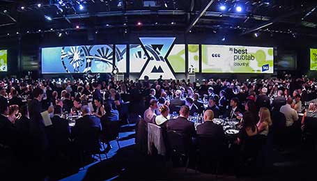 A large crowd dressed in formal attire sits at round tables in a spacious, dimly lit banquet hall, facing a stage set for a gala dinner, illuminated with screens displaying graphics and the words “best public sector” at an awards event.