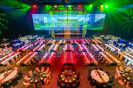 A large banquet hall is set up for an event, featuring long rectangular tables with colorful lighting in green, red, and yellow. Round tables line the edges, and a stage set for gala dinner with bright screens stands prominently at the front.