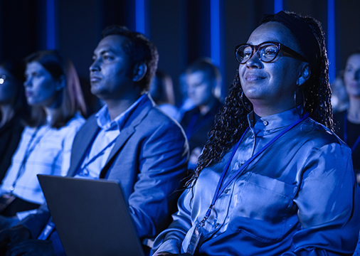 Audience members attentively watch a presentation in a dimly lit room enhanced by Event Technology. One person smiles while wearing glasses and a silk shirt, and another nearby has a laptop. The scene is illuminated with blue light.