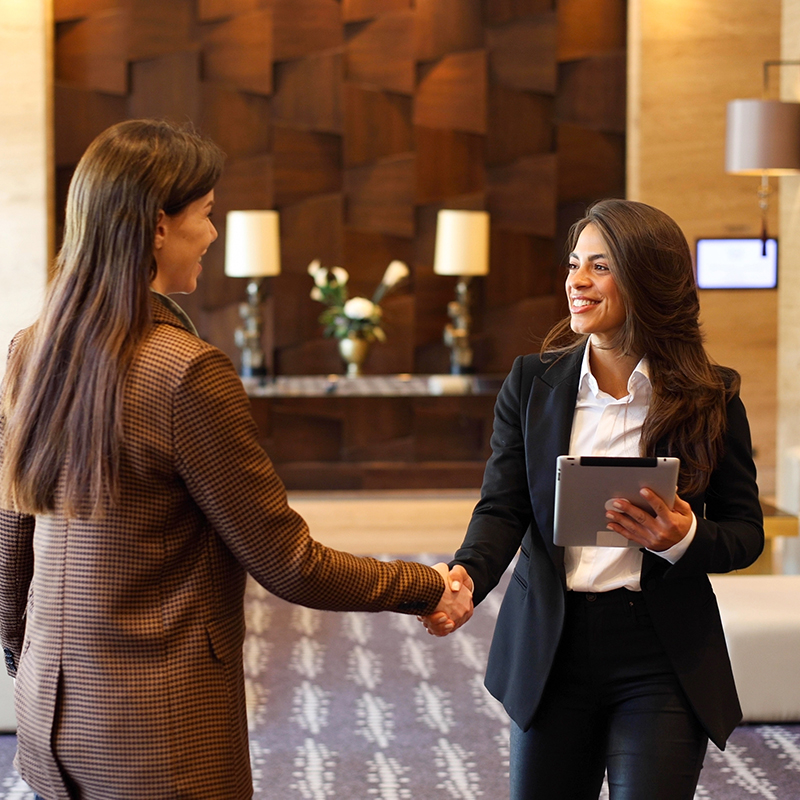 Two women in business attire shake hands and smile in a modern lobby with stylish decor, including lamps, flowers, and wood paneling. One holds a tablet.