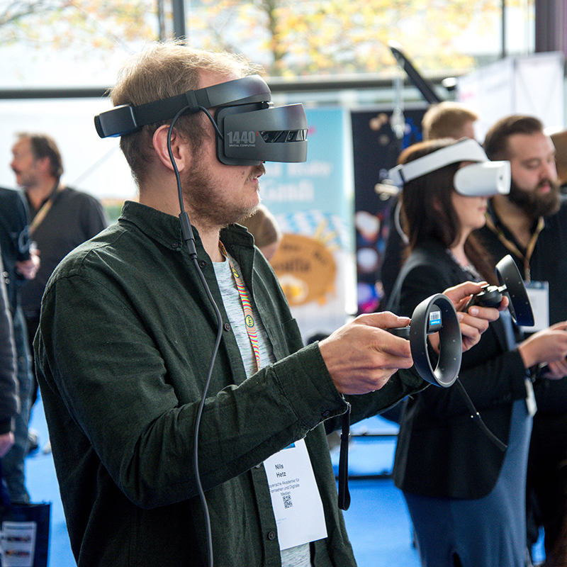 A man wearing a VR headset holds motion controllers at a tech event, surrounded by other people also using virtual reality equipment. The setting appears to be indoors with natural light coming through large windows.