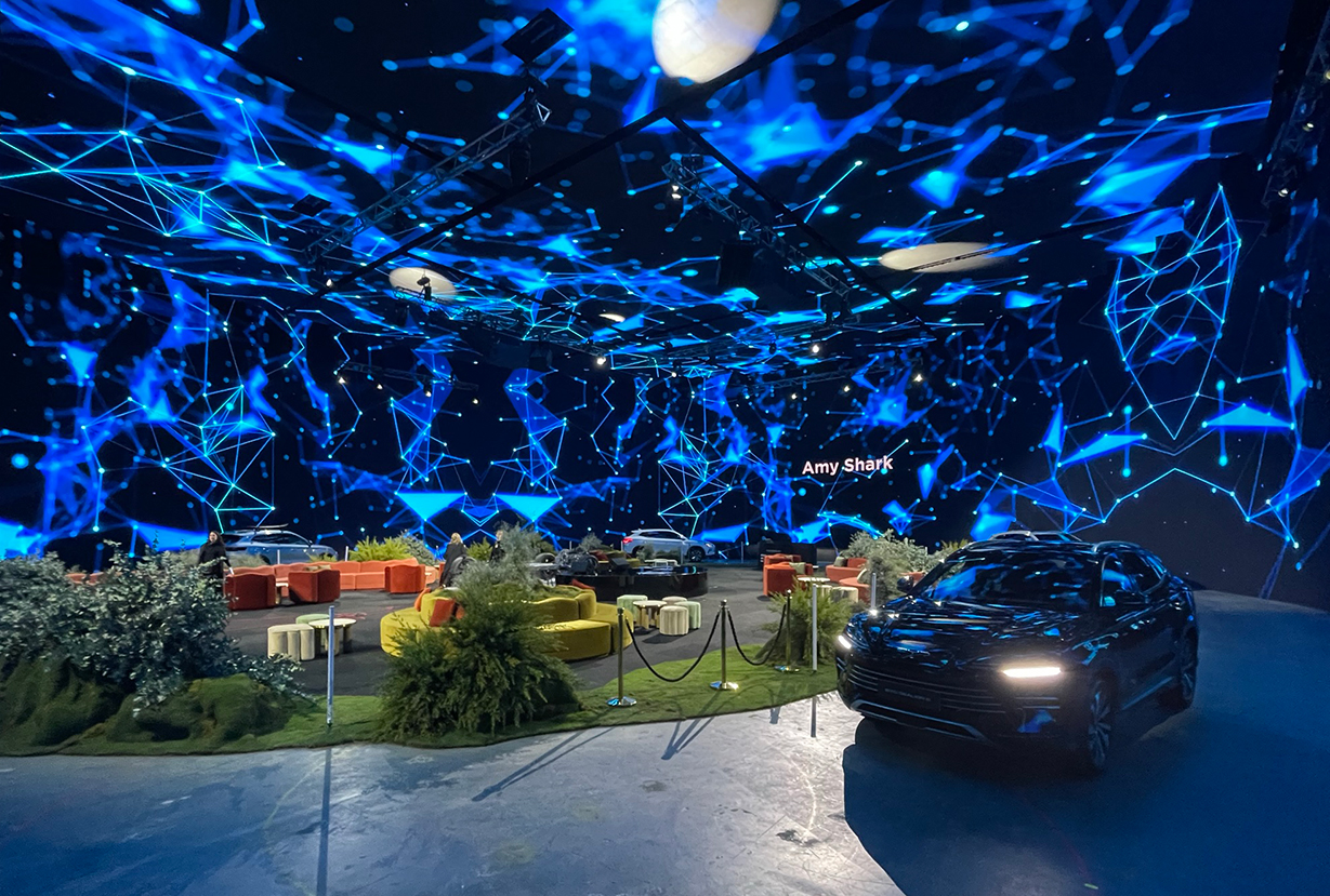 A futuristic indoor space with blue digital light projections, cutting-edge event technology, modern furniture, green plants, and a sleek black car in the foreground; a sign reads Amy Shark.