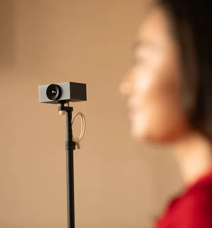 A small camera mounted on a tripod is in focus, while a person with long dark hair and wearing red is blurred in the background, facing the camera.