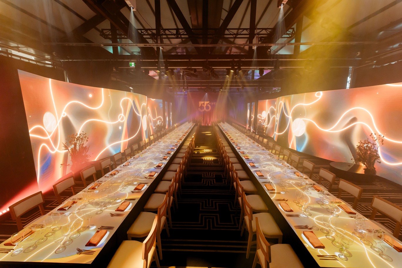 A long banquet table set for the Doltone 55 Years VIP Qantas Platinum Member Experience, with elegant place settings and centerpieces, flanked by illuminated, colorful abstract light panels under a high ceiling with dramatic lighting.