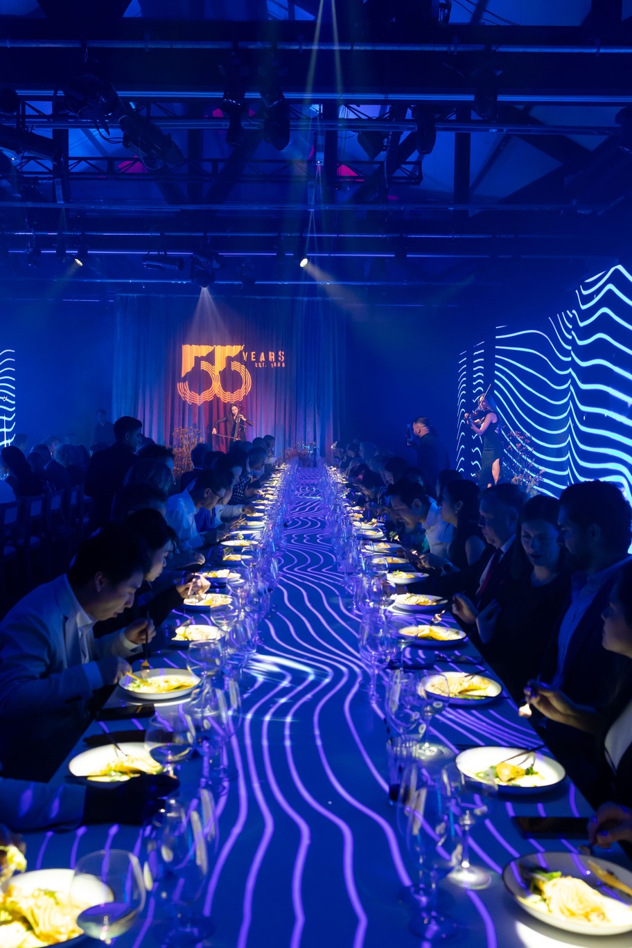 A long dinner table with guests seated on both sides is illuminated by blue and purple lights, with wavy light patterns projected onto the table. A 55 years sign glows on the stage at the end of the room.
